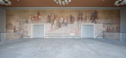 Photo of a mural: The painting depicts the visit to Dresden by the President of the GDR, Wilhelm Pieck, across the entire wall. He is surrounded by various people, some carrying flags. They are looking up at him or toward him.