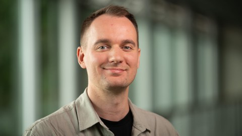 Poträtfoto: Aron Schulze lächelt in die Kamera. Er trägt eine Kurzhaarfrisur. Der Hintergrund ist verschwommen.