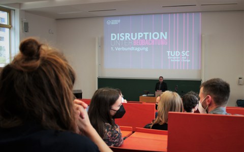 Blick in den Hörsaal von hinten. An der Wand eine Folie mit dem Titel "Disruption unter Beobachtung"