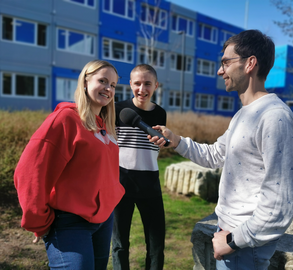Drei Personen vor dem Containerbau der Universitätsschule Dresden. Podcaster Fabian Deicke hält Schülerin Hedi das Mikro vors Gesicht. Sie lächelt in die Kamera. Im Hintergrund lacht Schüler Tjark in Hedis Richtung.