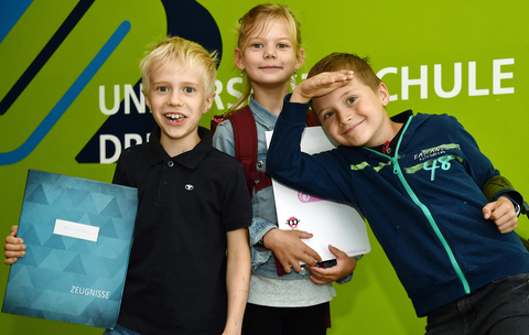 Drei Kinder stehen vor einer grünen Wand in der Universitätsschule Dresden und halten ihr Zeugnis lachend in die Kamera.