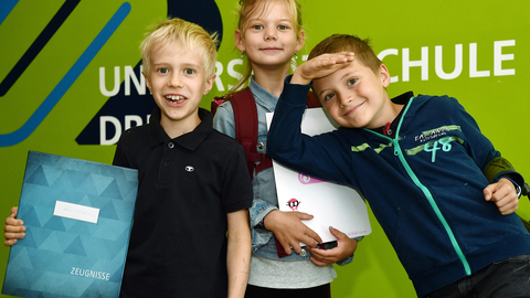 Drei Kinder stehen vor einer grünen Wand in der Universitätsschule Dresden und halten ihr Zeugnis lachend in die Kamera.