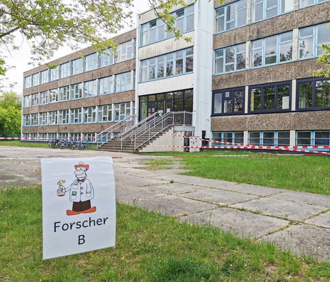 Das Unischulgebäude, ein DDR-Plattenbau, ist schräg von vorn zu sehen. Davor hängt an rot-weißem Flatterband ein Schild mit einer gezeichneten Comicfigur eines Forschers. Darunter steht "Gruppe Forscher B". Im Hintergrund ist der Schulhof mit rot-weißem Flatterband in verschiedene Wartezonen abgteilt. 