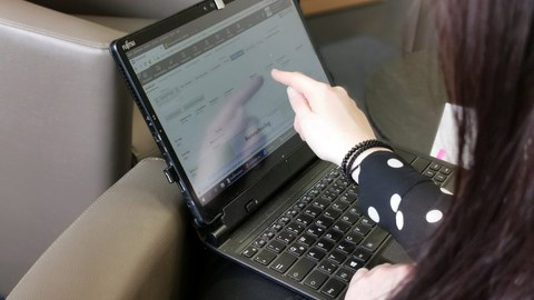 Eine Lehrerin erklärt am Laptop die Software der Universitätsschule Dresden fürs Homeschooling. Man sieht sie von hinten und ihre Hand zeigt auf den Bildschirm. 