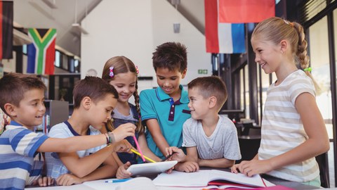 6 Kinder sitzen und stehen um einen Tisch in einer Schulbibliothek. Sie schauen auf ein Tablet.