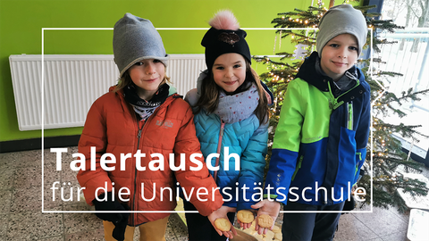 Drei Kinder in Winterjacken stehen im Foyer der Universitätsschule Dresden. Sie halten Plätzchen in Form des TU Dresden-Logos in die Kamera. Im Hintergrund steht ein beleuchteter Weihnachtsbaum.