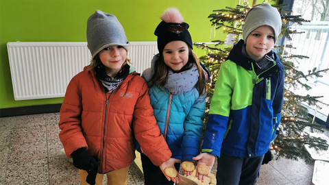 Drei Kinder in Winterjacken stehen im Foyer der Universitätsschule Dresden. Sie halten Plätzchen in Form des TU Dresden-Logos in die Kamera. Im Hintergrund steht ein beleuchteter Weihnachtsbaum.