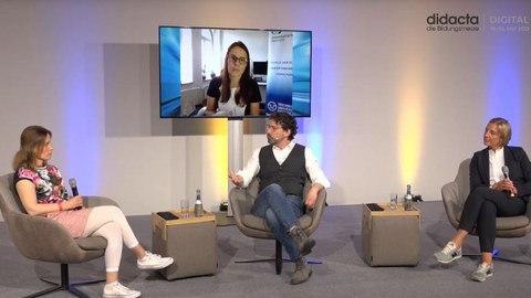 Bei einer Podiumsdiskussion sitzen zwei Schulleiterinnen und der Moderator gemeinsam im Studio. Auf einem großen Bildschirm ist Anke Langner, wissenschaftliche Leiterin der Universitätsschule Dresden zugeschaltet.