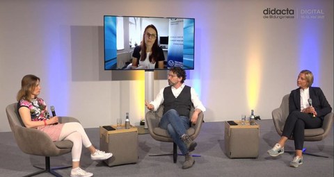 Bei einer Podiumsdiskussion sitzen zwei Schulleiterinnen und der Moderator gemeinsam im Studio. Auf einem großen Bildschirm ist Anke Langner, wissenschaftliche Leiterin der Universitätsschule Dresden zugeschaltet.