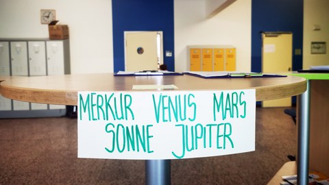An einem Stehtisch für die Corona-Anmeldung im Foyer der Universitätsschule Dresden klebt ein Zettel mit Stammgruppennamen als Aufschrift "Merkur Venus Mars Sonne Jupiter". Im Hintergrund sind blaue Lernraumtüren und orangefarbene Spinde.