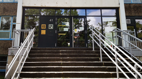 Frontale Ansicht des Haupteingangs der Universitätsschule Dresden. Eine Treppe führt in einen klassischen DDR-Schulplattenbau. In der Fensterfront spiegeln sich der blaue Sommerhimmel und die Baumkronen ringsum.