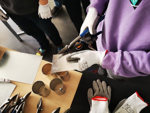 Blick von oben auf einen Werktisch mit Metallteilen. Zwei Hände mit Arbeitshandschuhen schneiden ein Blechstück zu.