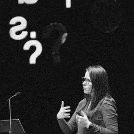Shcwarz-Weiß-Foto: Eine Frau steht in einem dunklen Raum und erklärt. Schräg über ihr hängen große weiße Buchstaben und Fragezeichen.