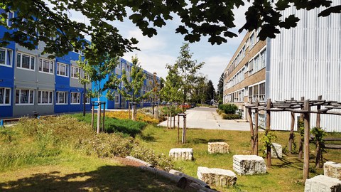 Das Schulgelände der Universitätsschule Dresden mit blau-grauem Containergebäude, DDR-Altbau und gestaltetem Außengelände als grünes Atelier im Sonnenschein