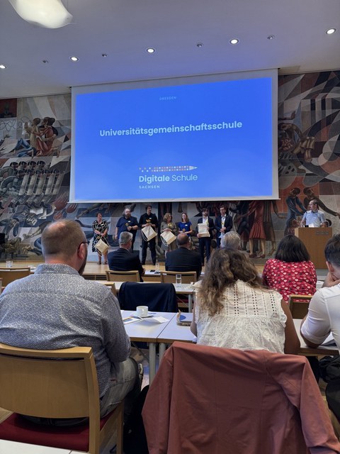 In einem Saal von hinten durch das Publikum aufgenommen der Titel "Universitätsgemeinschaftsschule" auf dem großen blauen Bildschirm. 