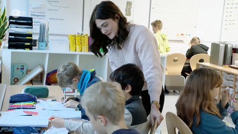 In einem Lernraum der Universitätsschule Dresden mit Regalen und Whiteboards sitzen drei Kinder an Arbeitstischen und schreiben. EIne junge Frau schaut ihnen über die Schulter.