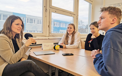 Eine Lernbegleiterin mit Mikrofon in der Hand, zwei Schülerinnen und ein Student sitzen für ein Interview um einen Arbeitstisch in der Unievrsitätsschule Dresden. Durch das Fenster sieht man im Hintergrund den Containerbau der Schule.