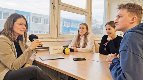 Eine Lernbegleiterin mit Mikrofon in der Hand, zwei Schülerinnen und ein Student sitzen für ein Interview um einen Arbeitstisch in der Unievrsitätsschule Dresden. Durch das Fenster sieht man im Hintergrund den Containerbau der Schule.