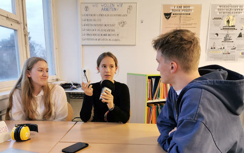 Zwei Schülerinnen und ein Student sitzen zum Interviewgespräch an einem Arbeitstisch in der Universitätsschule Dresden. Sie schauen sich an. Die Schülerin in der Mitte hält ein Mikrofon