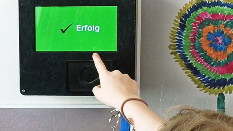 An einer Wand mit bunten Kinderbildern hängt ein schwarzer Kasten mit grünem Bildschirm. Auf diesem erscheint ein schwarzes Häkchen und der weiße Schriftzug "Erfolg". Eine Kinderhand hält einen Chip für die Anmeldung in der Hand.