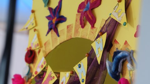 Gelbe Wimpelkette mit den Buchstaben WILLKOMMEN hängt an einer gelben Wand beklebt mit bunten Filzblumen