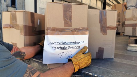 Zwei starke Arme heben Kartons mit Etikett "Universitätsgemeinschaftsschule" aus einem LKW.