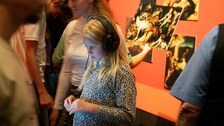 Ein Mädchen steht mit Kopfhörern in einem wuseligen Ausstellungsraum. Sie wird von einem Display angeleuchtet.