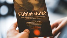 Eine Hand hält einen dunklen Flyer für die Ausstellungs "Fühlst Du's?"