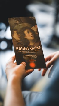 Eine Hand hält einen dunklen Flyer für die Ausstellungs "Fühlst Du's?"