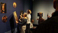 Vor einer dunkelblauen Ausstellungswand stehen zwei Frauen. Eine hält eins der abnehmbaren Bilder in der Hand.