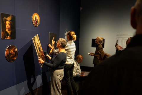 Vor einer dunkelblauen Ausstellungswand stehen zwei Frauen. Eine hält eins der abnehmbaren Bilder in der Hand.