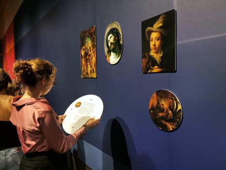 Vor einer dunkelblauen Ausstellungswand steht eine junge Frau. Sie liest den Text auf der Rückseite des abnehmbaren Bildes.