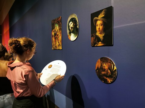 Vor einer dunkelblauen Ausstellungswand steht eine junge Frau. Sie liest den Text auf der Rückseite des abnehmbaren Bildes.