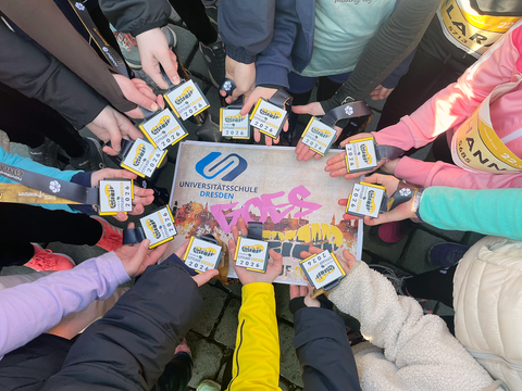 Um ein Schild mit der Aufschrift "Universitätsschule Dresden goes novaled Citylauf Dresden 2026" halten 12 Kinderhände eine Teilnehmerkarte für den Citylauf. Auf dem Schild sind die Silhouette der Stadt Dresden, ein Laufschuhabdruck und das USD-Logo