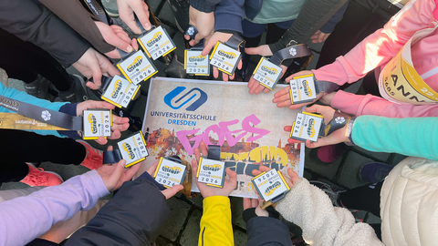 Um ein Schild mit der Aufschrift "Universitätsschule Dresden goes novaled Citylauf Dresden 2026" halten 12 Kinderhände eine Teilnehmerkarte für den Citylauf. Auf dem Schild sind die Silhouette der Stadt Dresden, ein Laufschuhabdruck und das USD-Logo