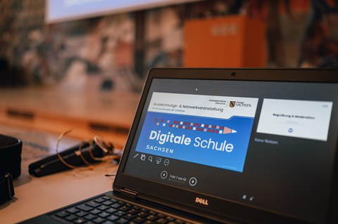 Logo und Schriftzug "Digitale Schule Sachsen" auf einem Laptopbildschrim, daneben ein Mikrofon, im Hintergrund ein Rednerpult vor der bunt gestalteten Wand im Saal der Dreikönigskirche Dresden