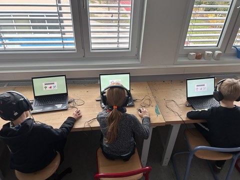 Drei Kinder sitzen mit Kopfhörern jeweils an einem Laptop mit der Comuptermaus in der Hand. Auf dem Bildschirm sind Texte und Bilder.