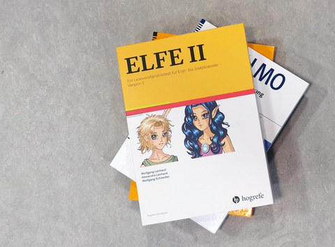 Auf einem Stapel liegen Ordner mit Unterhebungsmaterialien. Auf gelbem Grund der Titel "ELFE 2. Ein Leseverständnistest", darunter auf weißem Grund zwei Comicfiguren im Mangastil.