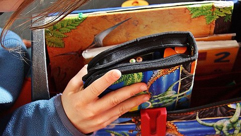 Eine Kinderhand hält eine bunte Federtasche vor einem Schulranzen.