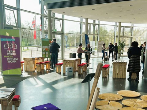 Etwa 10 Personen stehen in einem hellen Foyer mit Sitzgelegenheiten und Tischen neben dem Banner "Networking Lounge - Teamwork makes a dream work" 