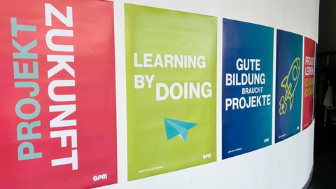 Eine Plakatwand mit den typographischen Motiven Zukunft, Learning by doing, Gute Bildung braucht Projekte! führt in einen Gang