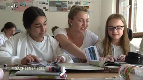 Screenshot des Beitrags aus dem MDR Sachsenspiegel vom Mittwoch, 5. Juni 2024. Eine Lehramtsstudentin schaut über die Schulter von zwei Schülerinnen und zeigt auf eine Stelle in einem Lehrbuch. 