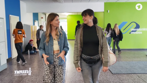Zwei junge Frauen laufen in einem Schulflur und unterhalten sich. Im Hintergrund eine grüne Wand mit dem Logo der Universitätsschule Dresden.