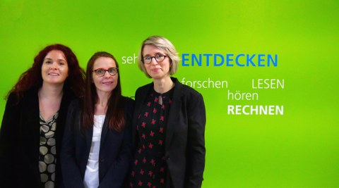 Vor einer leuchtend grünen Wand im Schulgebäude stehen drei Frauen, die Schulleiterinnen der Universitätsschule Dresden. Auf der Wand sind weißen und blauen Druckbuchstaben die Wörter entdecken, lesen, forschen, rechnen, hören geschrieben.