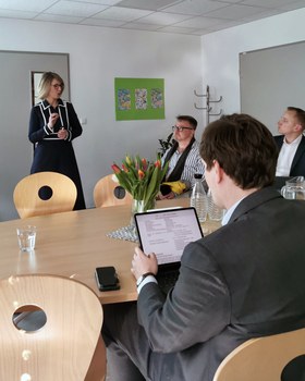 Schulleiterin Maxi Heß präsentiert in einem Besprechungsraum stehend vor drei sitzenden Männern. Kultusminister Clemens liest den Schulsteckbrief auf einem Tablet.