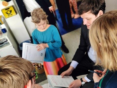 Drei Schüler:innen zeigen Kultusminister Conrad Clemens ihre Logbücher. Er sitzt auf einem Kinderstuhl und wird von ihnen umringt.