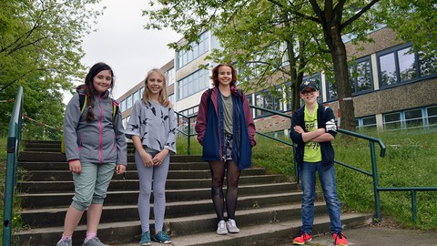 3 Schülerinnen und eine Studentin stehen auf der Treppe vor dem Schulgebäude