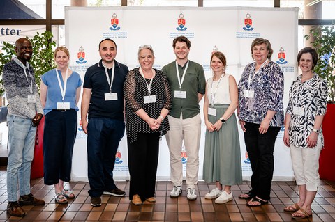 Acht Personen mit Konferenzschildchen stehen vor einer Fotowand mit dem Logo der Universität Pretoria