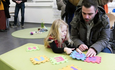 Ein Vater und seine Tochter knien auf dem Boden hinter einem grünen Sofa in einem Lernraum. Auf der Sitzfläche puzzeln sie aus großen Buchstaben S C H U L E das Wort Schule. Im Hintergrund stehen Eltern und Lernbegleiter im Informationsgespräch.