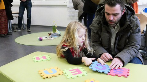 Ein Vater und seine Tochter knien auf dem Boden hinter einem grünen Sofa in einem Lernraum. Auf der Sitzfläche puzzeln sie aus großen Buchstaben S C H U L E das Wort Schule. Im Hintergrund stehen Eltern und Lernbegleiter im Informationsgespräch.